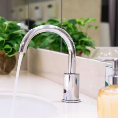 A modern bathroom faucet with water running into a white sink. On the countertop is a soap dispenser filled with liquid soap. In the background, green plants add a touch of nature to the clean, reflective bathroom setting.