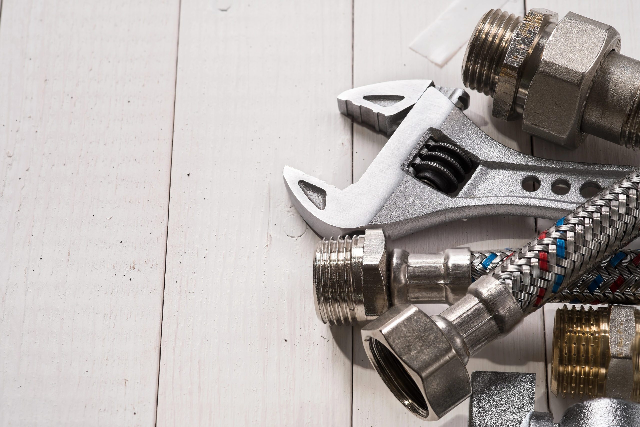 A close-up of various plumbing supplies arranged on a light wooden surface, including a wrench, metal fittings, and braided hoses. The items are positioned on the left side of the image, while the right side shows the plain wooden texture.