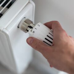 A close-up of a hand adjusting a white thermostat on a radiator. The thermostat is marked with numbers and symbols, suggesting different heat settings. The radiator appears clean and is installed against a light-colored wall.