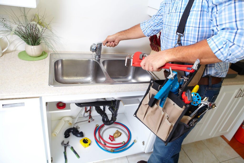 A plumber wearing a blue checkered shirt is fixing a kitchen sink with a red pipe wrench. He has a tool bag attached to his waist filled with various tools. The under-sink cabinet door is open, revealing plumbing supplies and equipment.