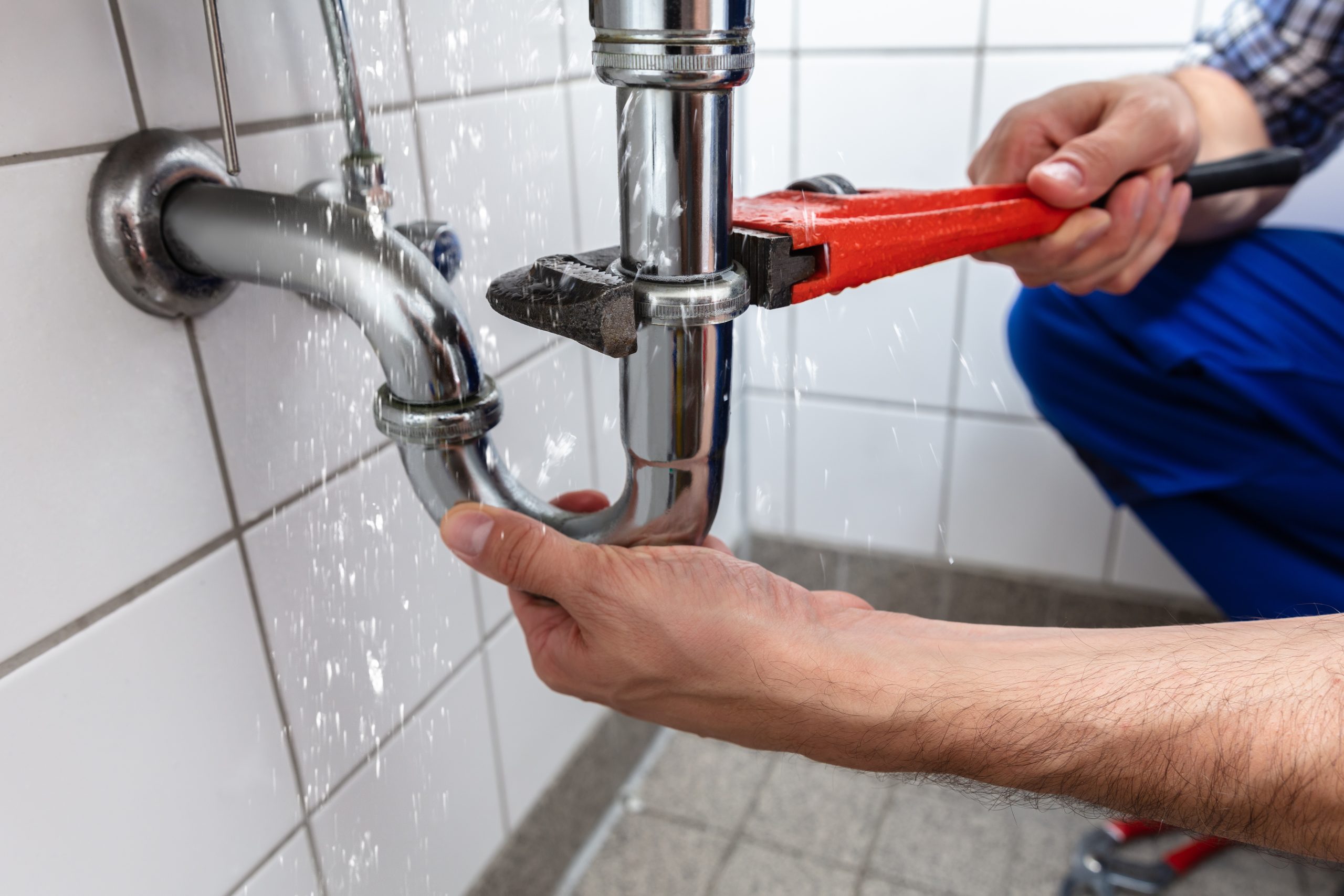 A person using a red wrench to fix a leaking pipe under a sink. Water is spraying from the pipe, and the person's hands are holding the pipe and the wrench, attempting to stop the leak. The background shows white tiled walls and a gray tiled floor.