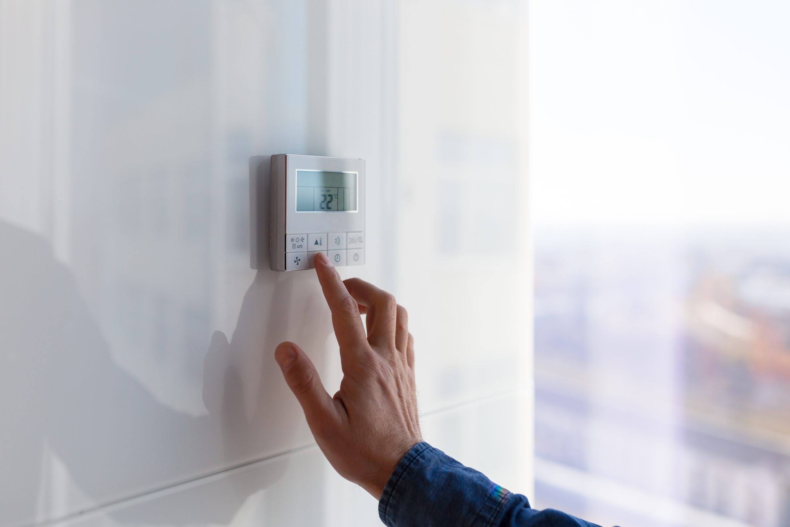 A person adjusting the settings on a digital thermostat mounted on a white wall. A finger is pressing one of the buttons, and the screen displays a temperature of 22°C. The background is blurred, showing a bright room with sunlight streaming in.