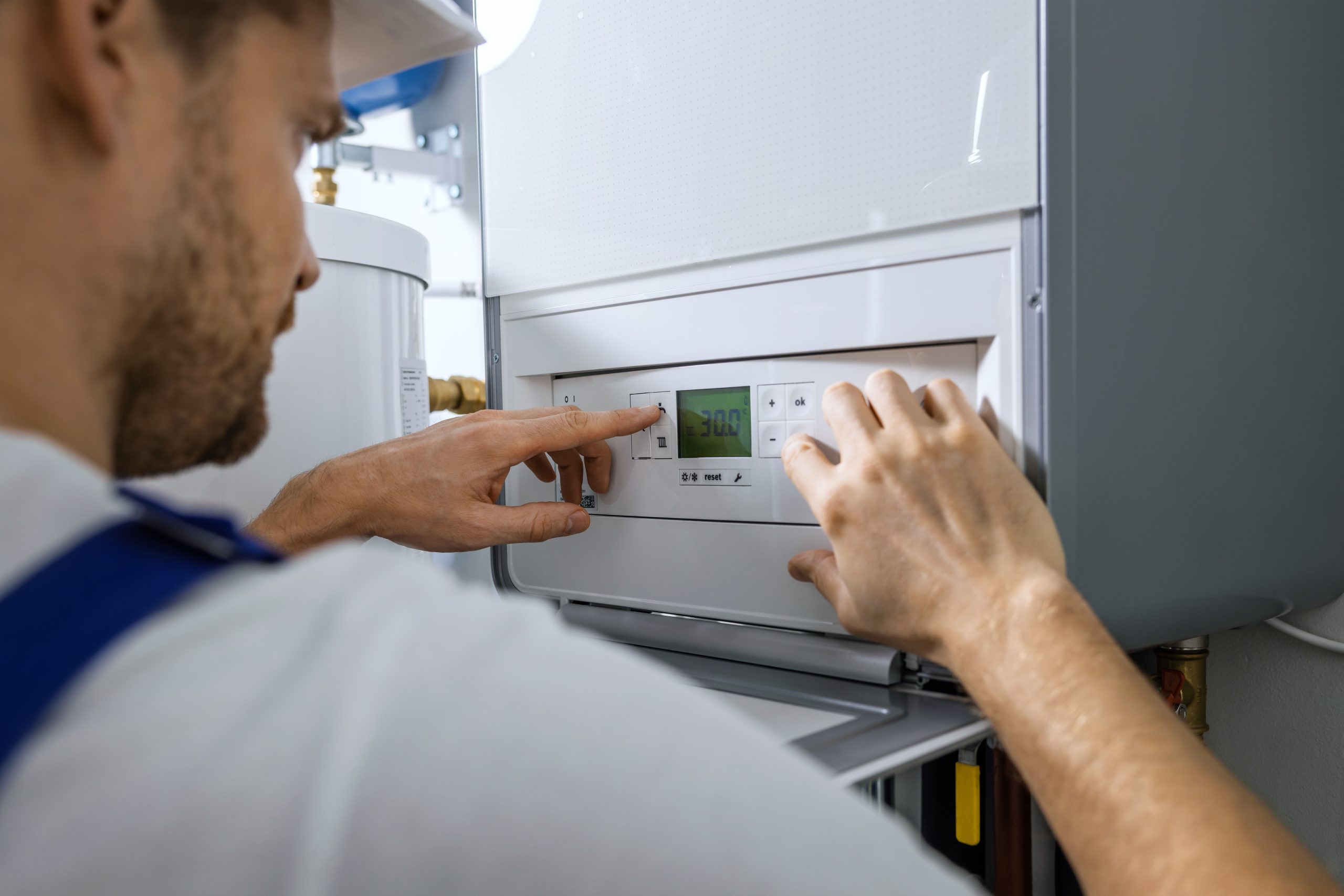 A technician wearing a hard hat adjusts the settings on a control panel of a heating system. His hands are seen turning a dial and pressing buttons. The panel displays a digital screen showing the number "30.0". The background includes part of the system's machinery.