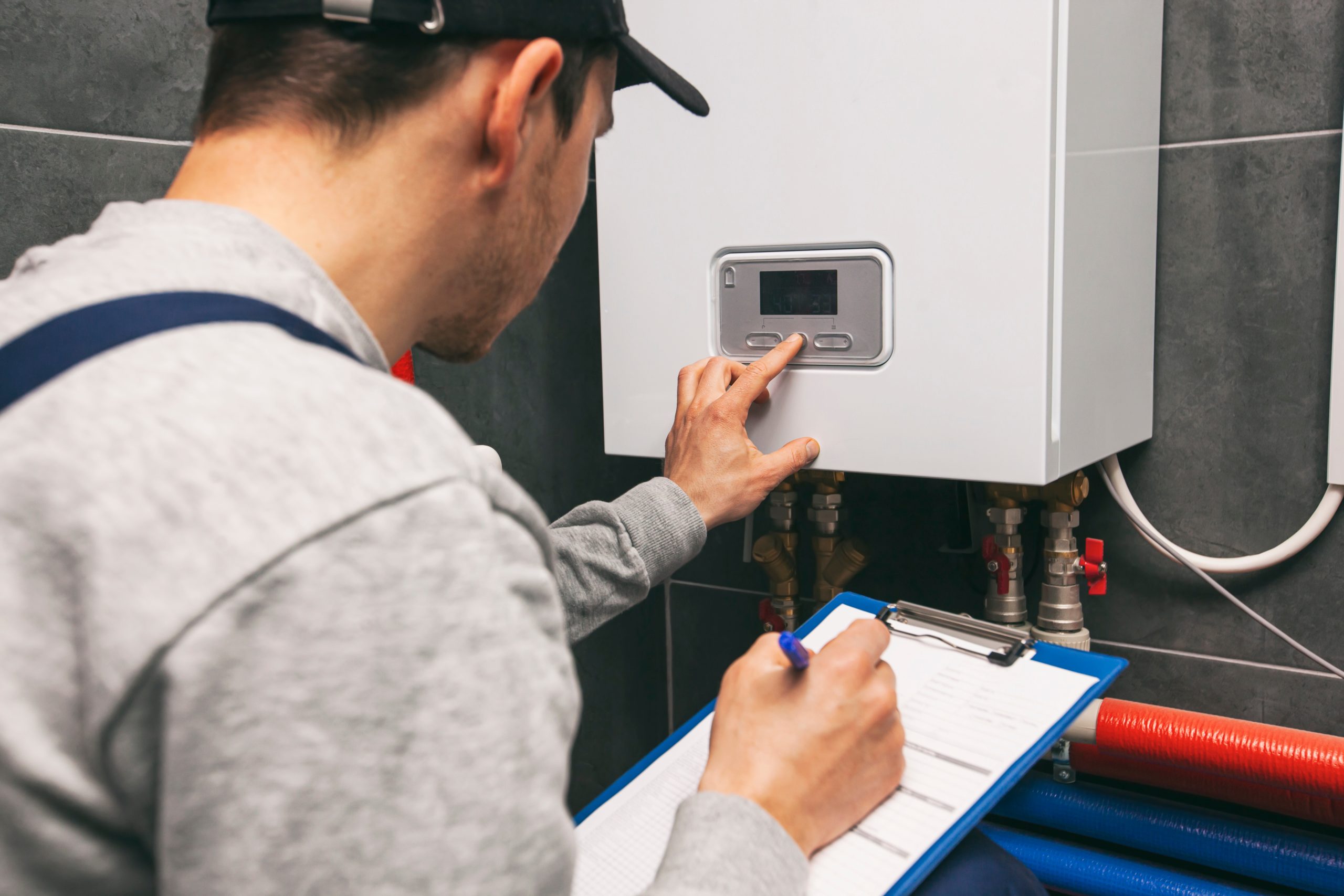 A person in a cap and gray sweatshirt adjusts the settings on a wall-mounted boiler while holding a clipboard and pen. Blue and red pipes are visible below the boiler. The scene appears to be in a utility or maintenance room.