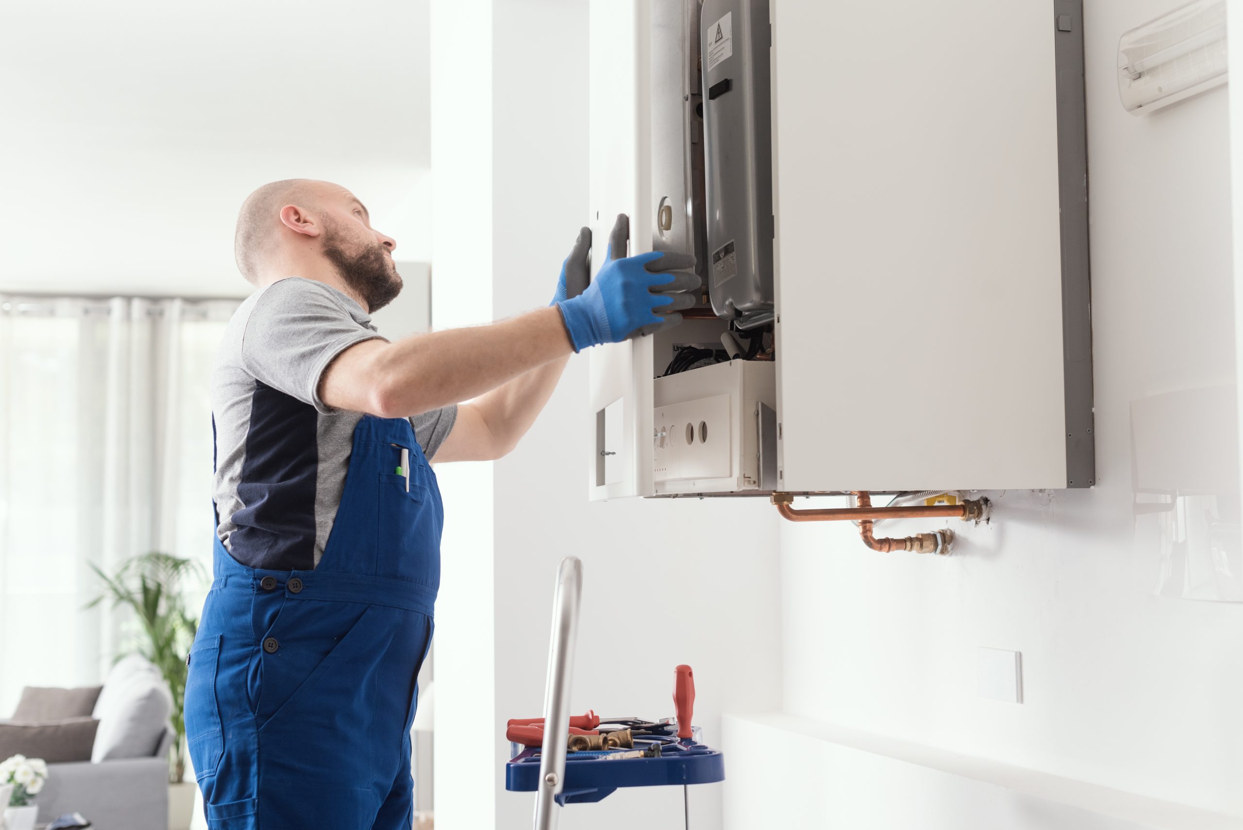 A technician in blue overalls and gloves is working on a wall-mounted boiler in a modern home. Tools are set on a nearby ladder in a well-lit room with a sofa and plant partially visible in the background.