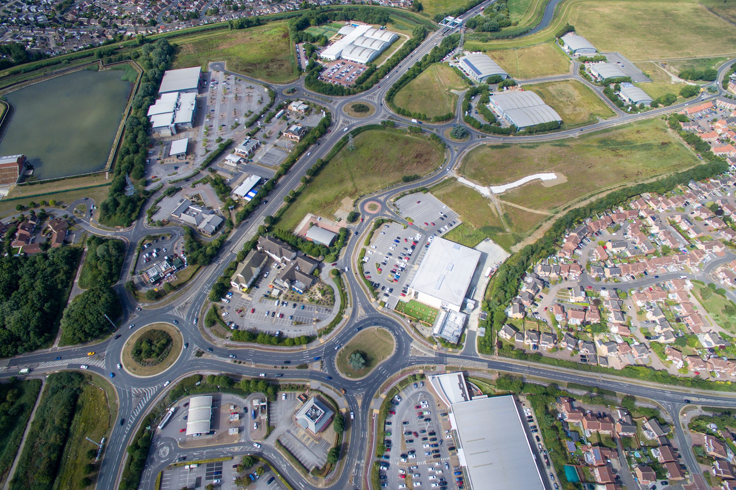 Aerial view of a suburban area featuring roundabouts, residential homes, and commercial buildings. Roads connect various sections, with green spaces and parking lots scattered throughout. The layout showcases a mix of housing and industrial structures.