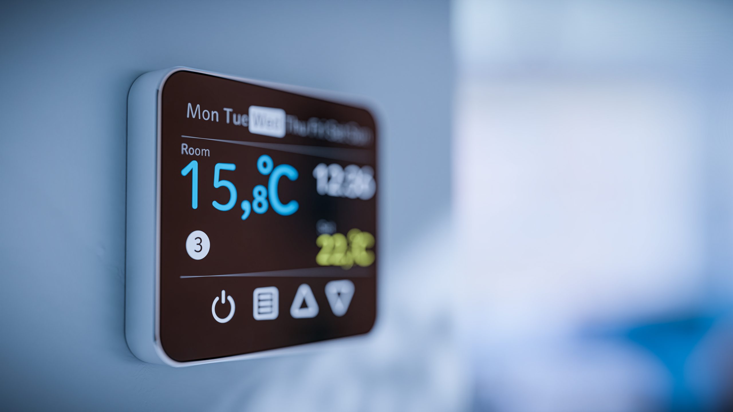 A close-up of a digital thermostat mounted on a wall displaying a room temperature of 15.8°C. The screen shows the current day as Tuesday, the time as 12:36, and other control icons. The background is out of focus, creating a modern, minimalistic look.